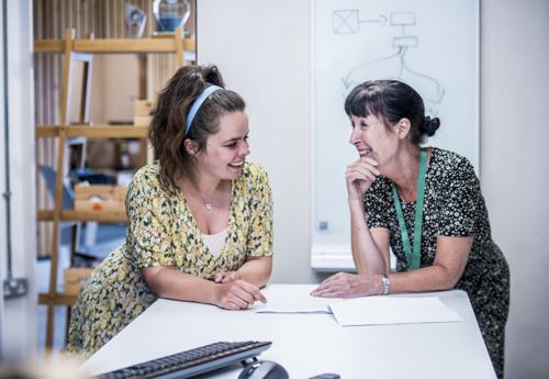 Income Manager Sharron and Head of Neighbourhoods Steff, stand together smiling and talking as they lean on an office desk