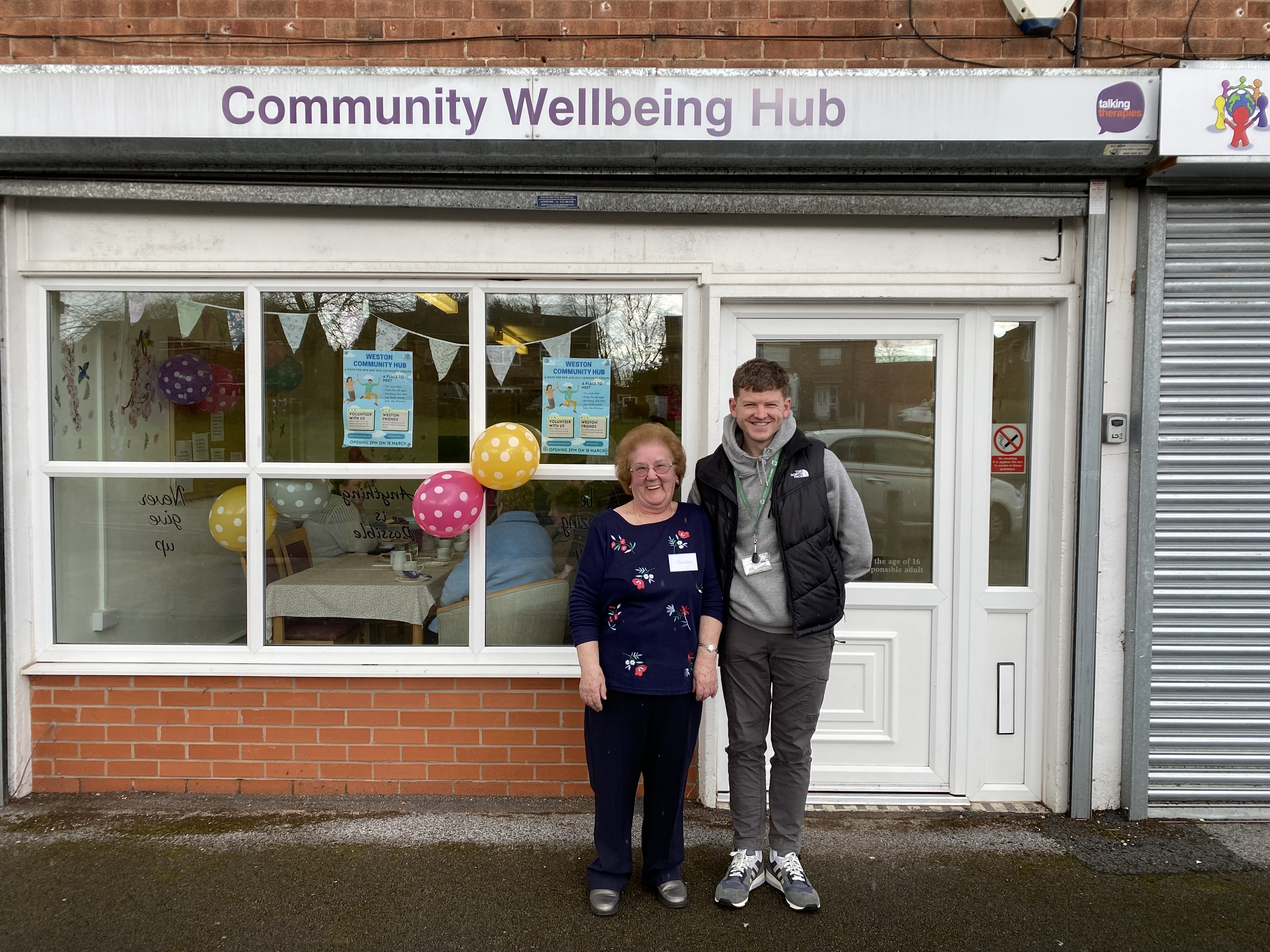 Two people stand outside the Weston Community Hub a sign reads Community Wellbeing Hub
