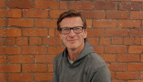 Man in spectacles and a grey hoodie in front of a brick wall