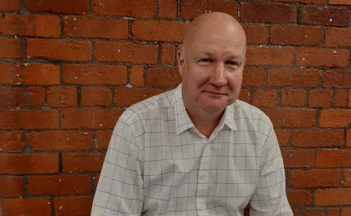 Man in white check shirt in front of a brick wall
